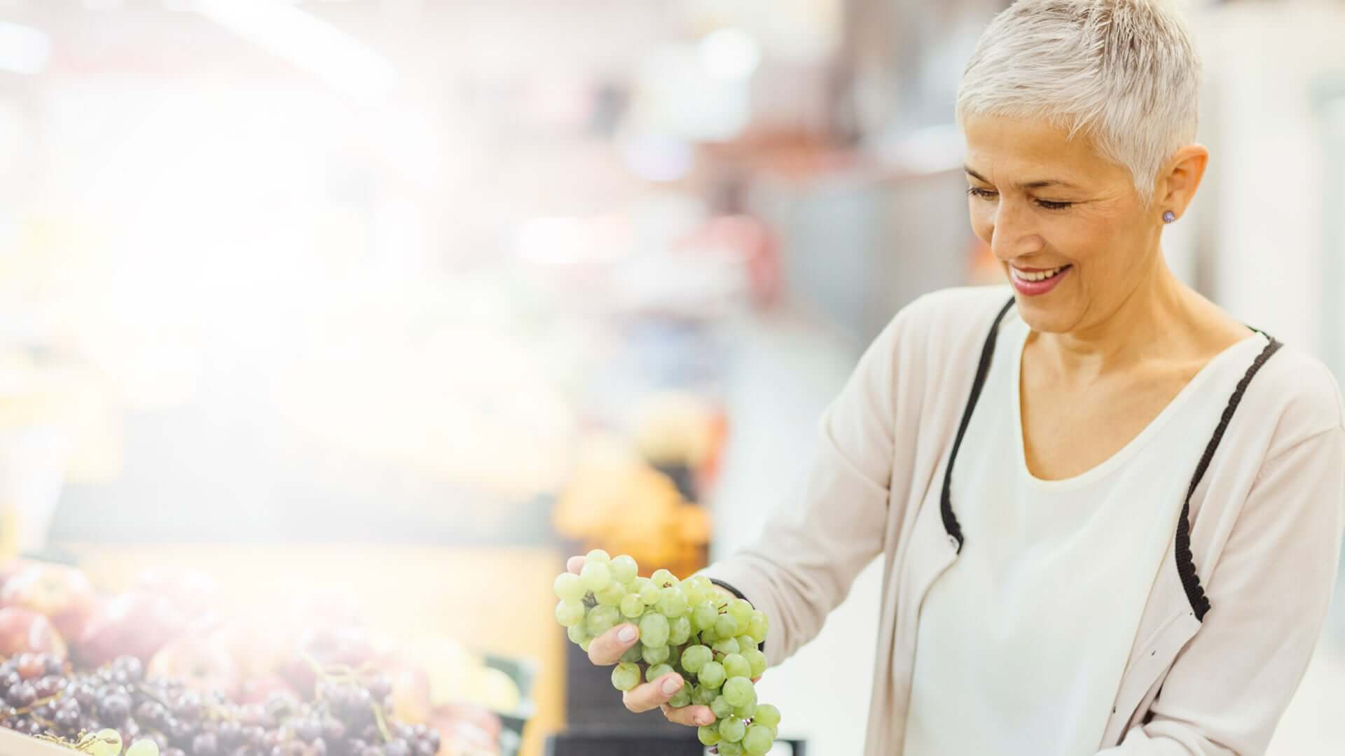 Mature smiling woman shopping in local supermarket. She is shopping groceries, choosing fruits, grapes.