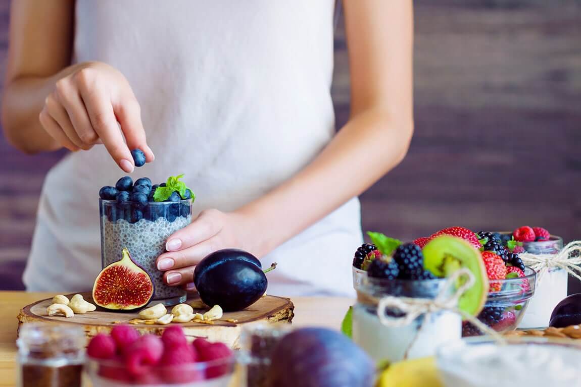 Female hands are preparing yogurt with chia and blueberries for good digestion, functioning of gastrointestinal tract. Summer berries, nuts, fruits, dairy products on table.