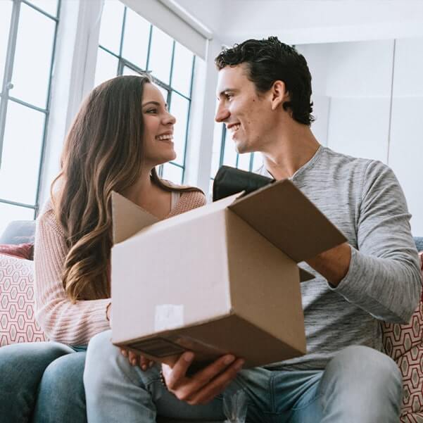 natural-health-strategies-rbti-testing-04-virtual-appt-final-min A cheerful couple sits in their home living room opening a cardboard box package from an online delivery