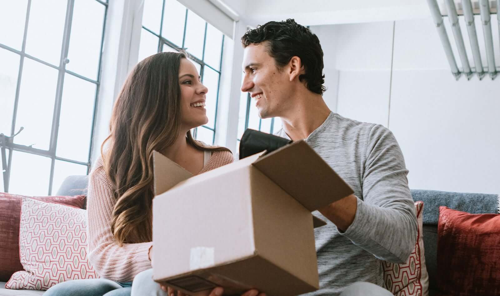 A cheerful couple sits in their home living room opening a cardboard box package from an online delivery